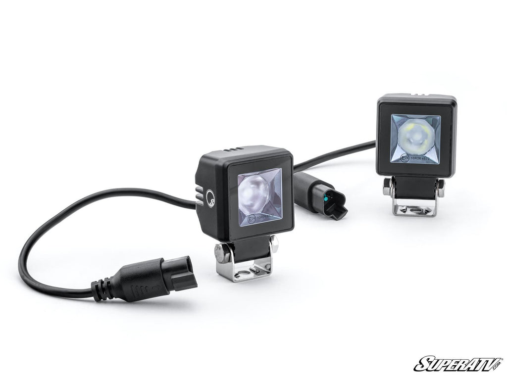 Two CFMOTO UForce 1000 Rear-Facing LED Cube Lights with clear lenses are wired together and mounted on silver brackets. One faces forward, the other is angled. A close-up highlights power connectors. The SuperATV logo appears bottom right on white.