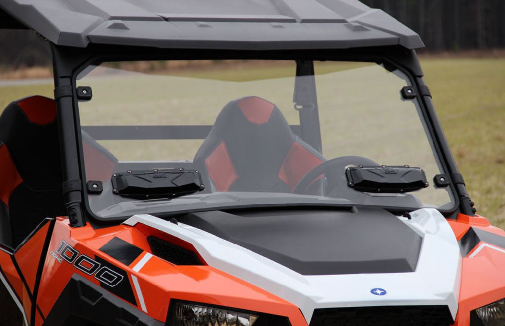 A close-up of a Polaris General with the Windshield Versa-Vent (Uncoated Poly) installed, featuring large vents, geometric-patterned seats, and two black dashboard devices. Grassy fields and trees appear in the cloudy background.