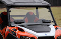 A close-up of a Polaris General with the Windshield Versa-Vent (Uncoated Poly) installed, featuring large vents, geometric-patterned seats, and two black dashboard devices. Grassy fields and trees appear in the cloudy background.