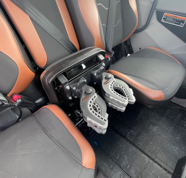 The Offroad Armor Can-Am Defender Hidden Rifle Mount is installed in a car interior with black and brown leather seats. The middle seatback folds down to reveal two rugged, mechanical plastic clamps where the seat normally sits.