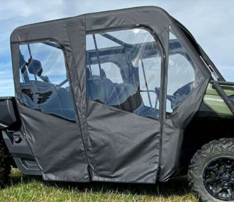 A side view of a Can Am Defender Max with Full Soft Doors, featuring black fabric and clear plastic windows that show the interior seats. The vehicle sits on grass under a blue sky, with Defender visible on the front fender.