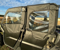Close-up of a black off-road vehicle parked on grass by a field, equipped with Can-Am Defender Max Soft Upper Doors For Lower Doors. Sunlit clear plastic windows and fabric doors are visible; the sky is partly cloudy, with a wooden fence in the background
