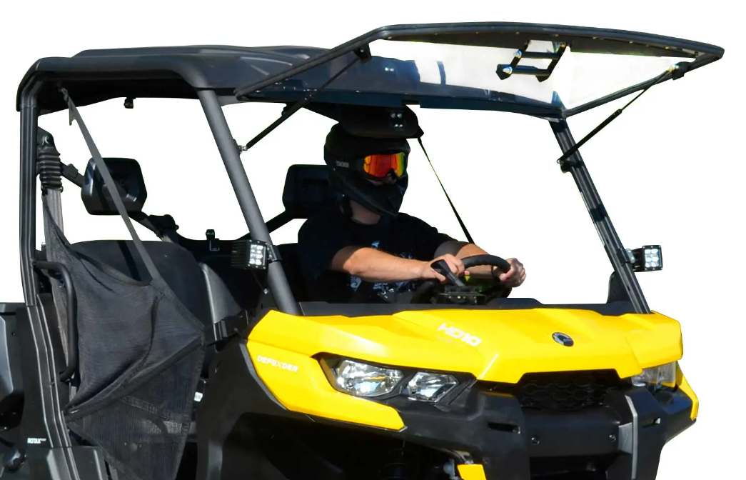 A person wearing a helmet and goggles drives an off-road vehicle equipped with the Can-Am Defender Scratch Resistant Flip Windshield, featuring mesh doors, a roof, headlights, and gripping the steering wheel while looking ahead.