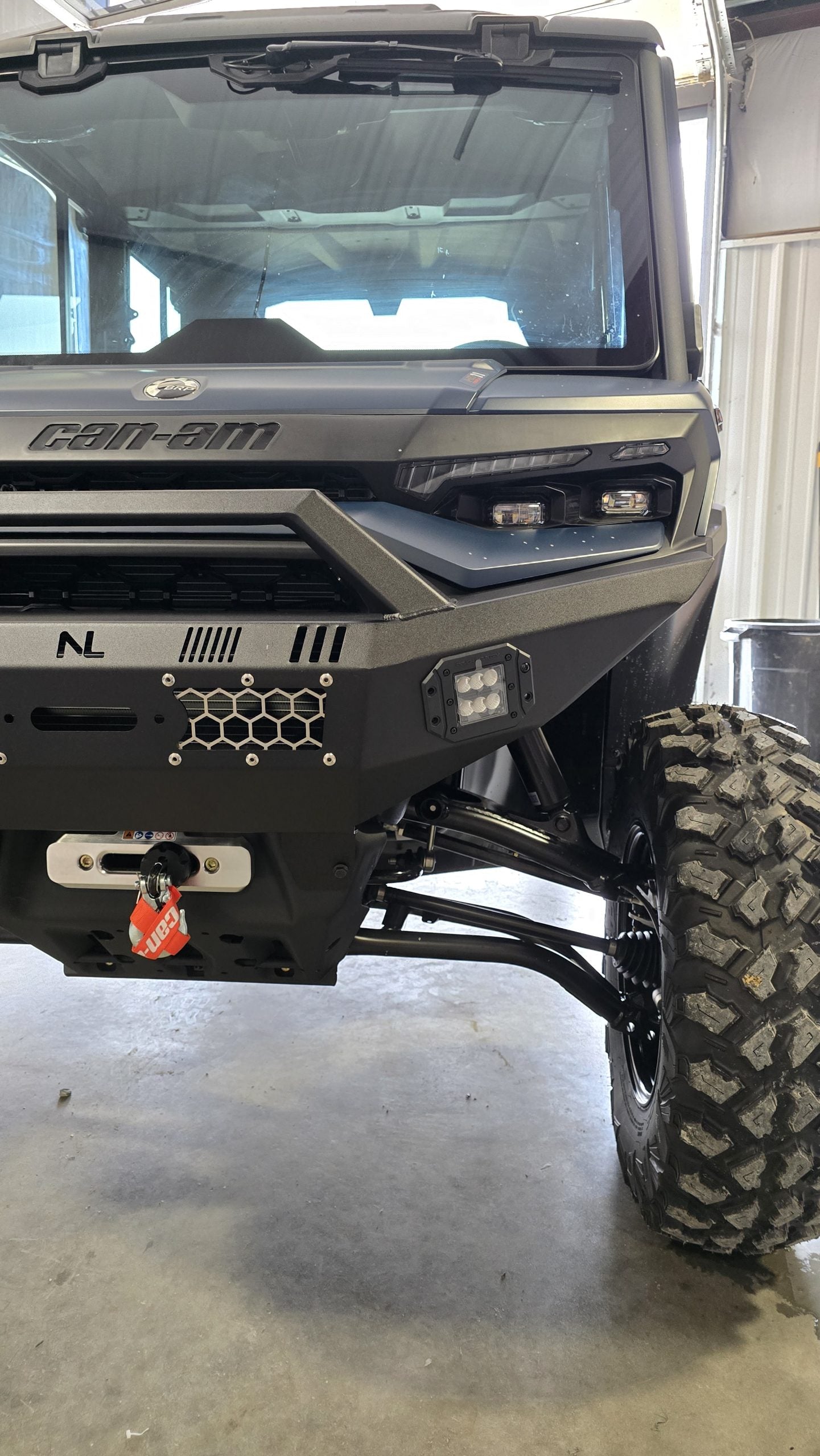 Close-up of the 2026 Can-am Defender HD11 Front Winch Bumper with lights, showing its rugged honeycomb design, winch with red strap, large treaded tire, and partially visible LED light inside a garage.