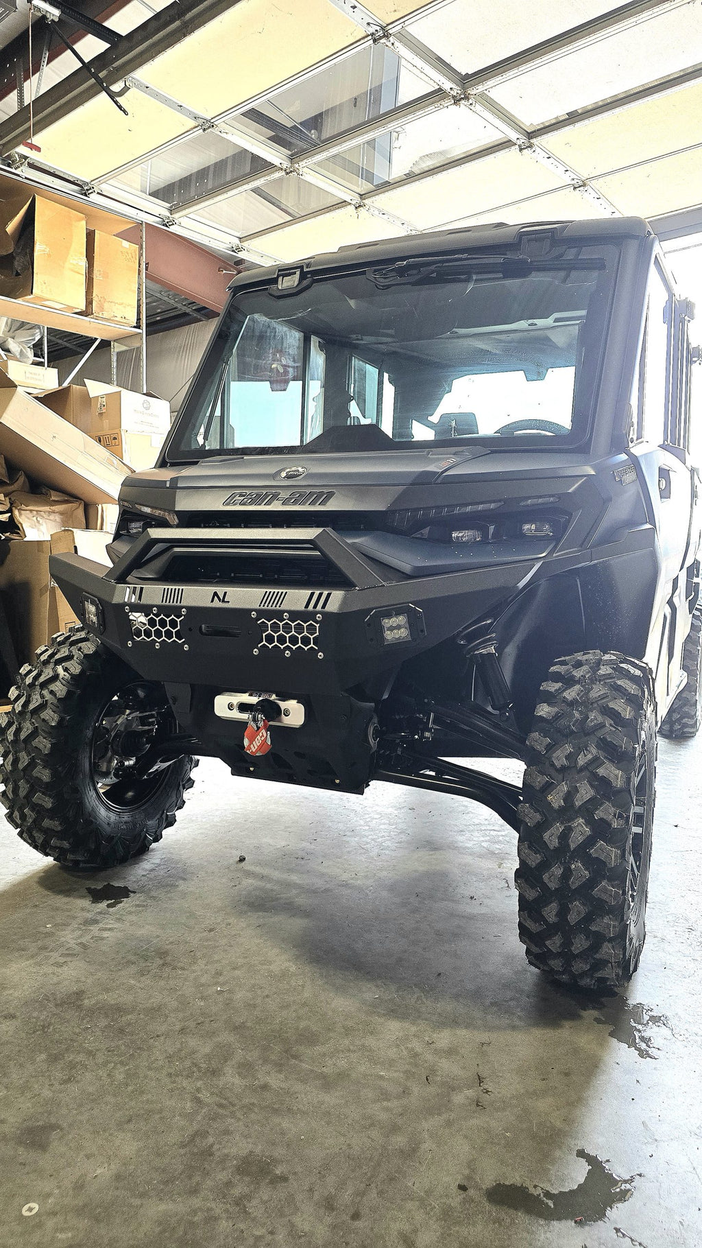 A 2026 Can-am Defender HD11 with a front winch bumper and lights is parked in a garage filled with boxes and equipment. The open garage door lets in natural light, highlighting the vehicle’s rugged tires, bumper, and grille.
