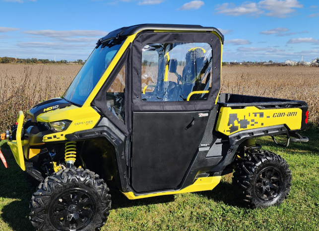 A Can-Am Defender with Convertible Doors (2016-Current, all models) in yellow and black is parked on grass under a blue sky. It features large off-road tires, a covered cab with clear windows, and bold black and yellow accents.