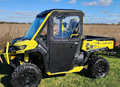 A Can-Am Defender with Convertible Doors (2016-Current, all models) in yellow and black is parked on grass under a blue sky. It features large off-road tires, a covered cab with clear windows, and bold black and yellow accents.