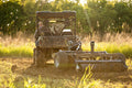 A person operates a utility vehicle towing the Heavy Duty Commercial Grade Disc/Culti-Packer Combo across a sunlit grassy field, turning fresh soil as warm sunlight filters through green trees in the background.