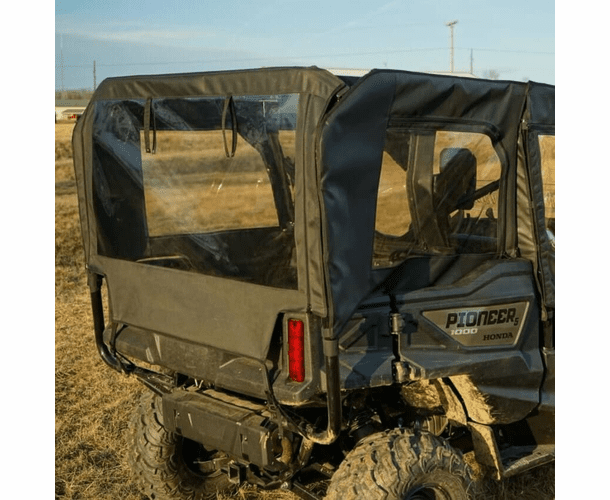 A black Honda Pioneer 1000 outfitted with Honda Pioneer Soft Upper Rear Doors, Middle and Rear Windows is parked on dry grass. The clear enclosure covers the cab for interior protection. The background features an open, flat landscape under a clear sky.