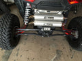 Close-up of Assault Industries High Clearance Quick Camber Radius Rods installed on a Polaris RZR XP 1000s rear suspension, with rugged tires, dual Muzzy exhaust, coilover shocks, and an LED light on the axle housing above a concrete floor.