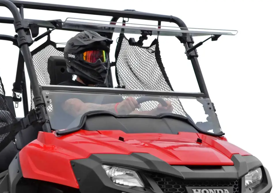 A person in a black helmet and goggles drives a red Honda off-road vehicle with a Honda Pioneer 700 Scratch Resistant Flip Windshield, mesh safety net, windshield bar, rugged design, and visible Honda logo. The background is plain white.