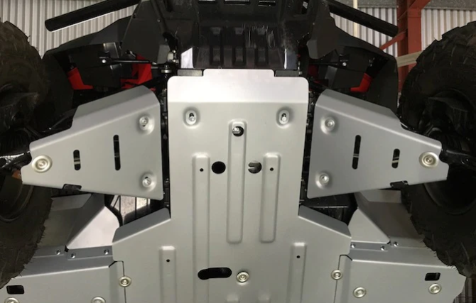 A close-up of the underside of a Honda Pioneer 1000 (2022+) shows silver alloy front A Arm Guards bolted in place, protecting the chassis. Rugged tires are visible on either side, with some framework above.