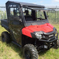 A Honda Pioneer 700 (2014+) with the Full Hard Cab Enclosure by Hard Cabs, featuring a roof, windshield, and side doors, sits on grass near a metal fence. The rugged utility vehicle is built for off-road work under a partly cloudy sky.
