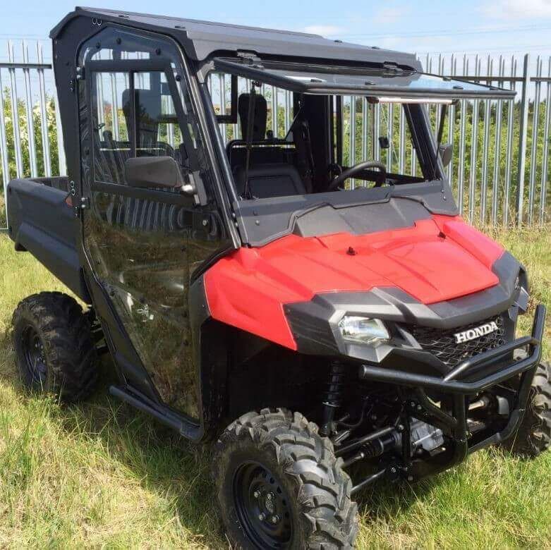A Honda Pioneer 700 (2014+) with the Full Hard Cab Enclosure by Hard Cabs, featuring a roof, windshield, and side doors, sits on grass near a metal fence. The rugged utility vehicle is built for off-road work under a partly cloudy sky.
