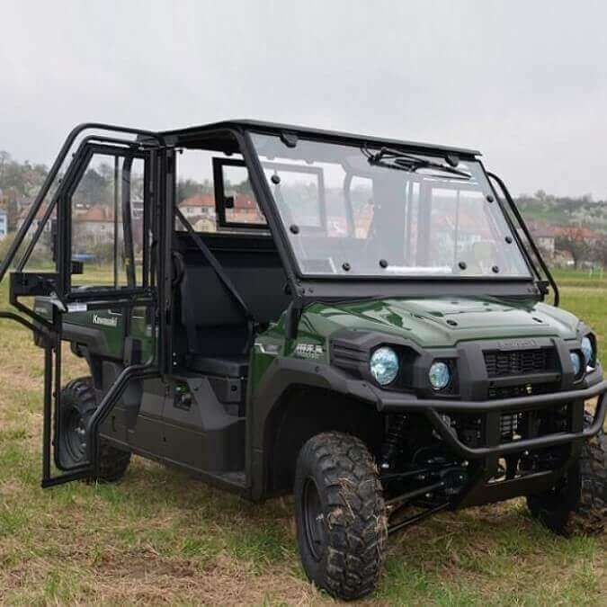 A Full Hard Cab Enclosure by Hard Cabs for a 2016-21 Kawasaki Mule Pro-Fx | Dx UTV, in green with black frames and rugged tires, is parked on a grassy field with open doors and windows. Trees and blurred buildings appear in the background under an overcas