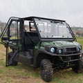 A Full Hard Cab Enclosure by Hard Cabs for a 2016-21 Kawasaki Mule Pro-Fx | Dx UTV, in green with black frames and rugged tires, is parked on a grassy field with open doors and windows. Trees and blurred buildings appear in the background under an overcas