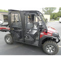 A 2015-21 Kawasaki Mule Pro-Fxt with a Full Hard Cab Enclosure by Hard Cabs, featuring a closed cabin, large off-road tires, two-row seating, barred side windows, and rear cargo bed, parked outside industrial buildings on a cloudy day.