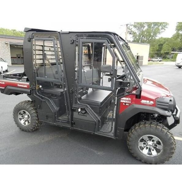 A 2015-21 Kawasaki Mule Pro-Fxt with a Full Hard Cab Enclosure by Hard Cabs, featuring a closed cabin, large off-road tires, two-row seating, barred side windows, and rear cargo bed, parked outside industrial buildings on a cloudy day.