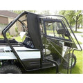 A close-up side view of a white 2016-19 Kawasaki Teryx 800 with the Full Hard Cab Enclosure by Hard Cabs, featuring a transparent angular door and window. The vehicle is parked on grass near houses and trees; the sturdy door frame is black.