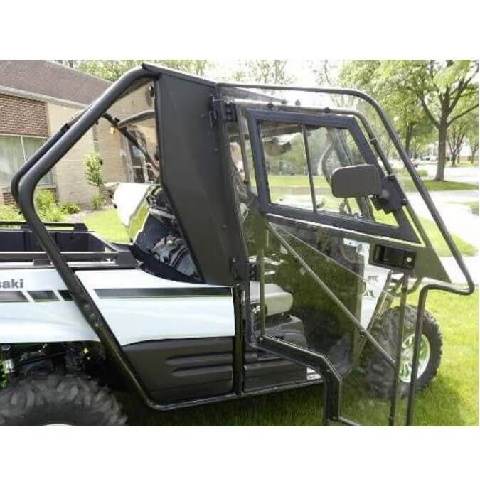 A close-up side view of a white 2016-19 Kawasaki Teryx 800 with the Full Hard Cab Enclosure by Hard Cabs, featuring a transparent angular door and window. The vehicle is parked on grass near houses and trees; the sturdy door frame is black.