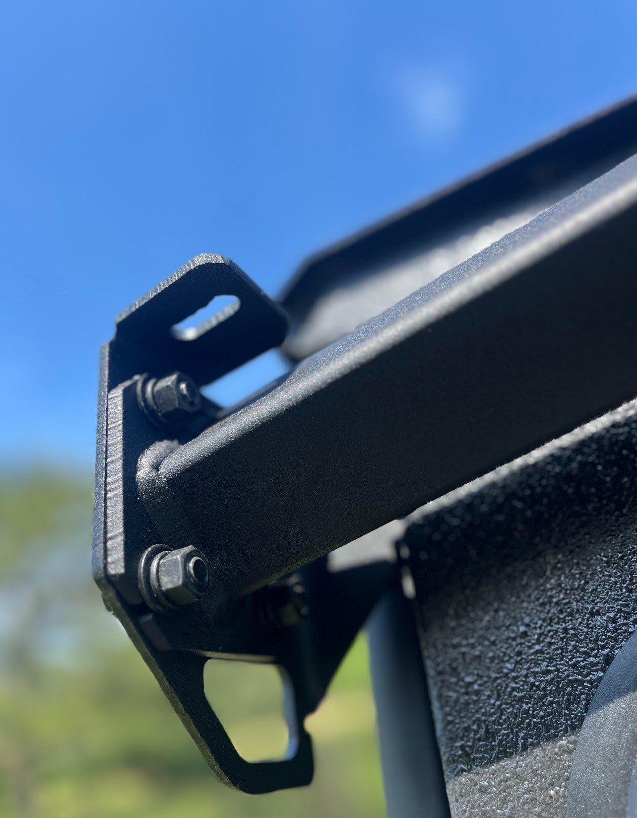 Close-up of the Defender Game Loader’s black metal bracket, bolted to a textured black surface. The blurred background reveals blue sky and greenery, emphasizing the loaders durable hardware and robust outdoor-ready construction.
