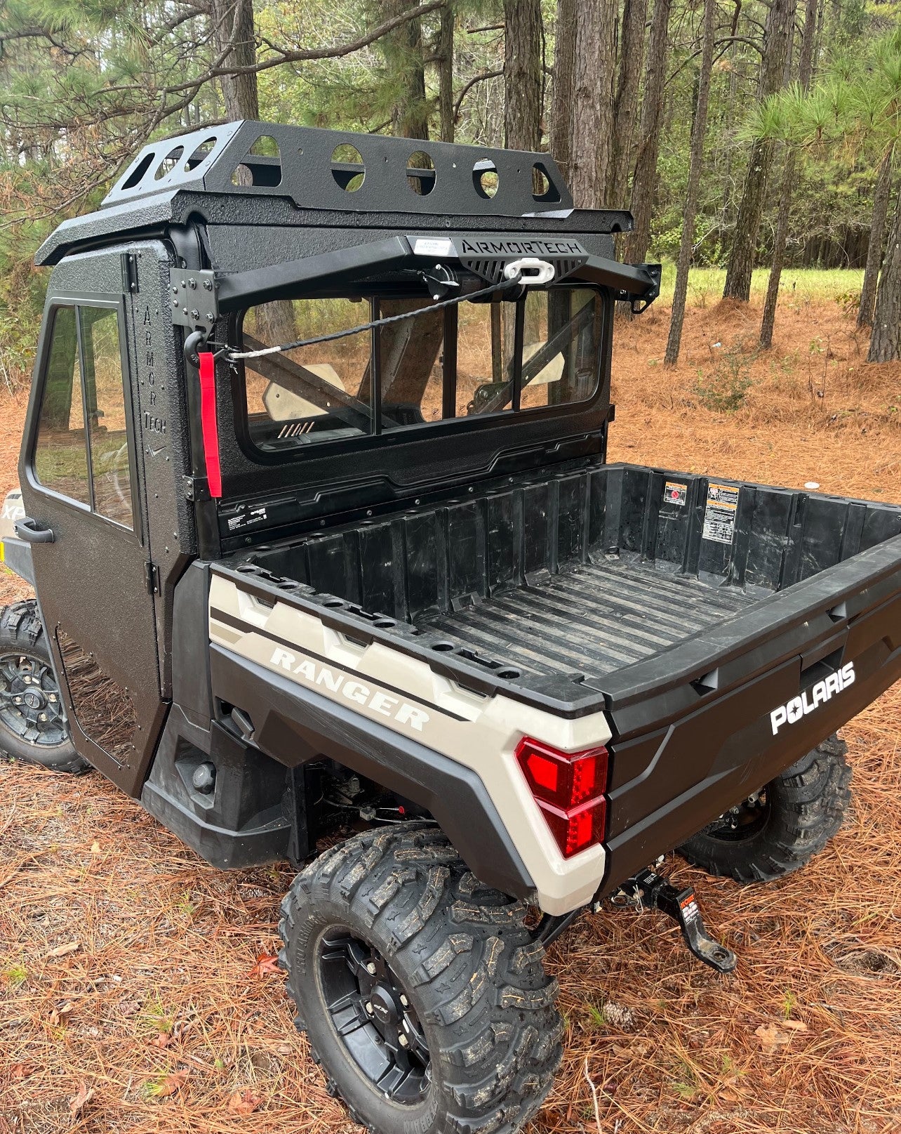 A black and tan Defender Game Loader utility vehicle with rugged tires and a metal roof rack is parked on a pine needle-covered forest floor. Its rear cargo bed is empty, and green-needled pine trees surround the area.