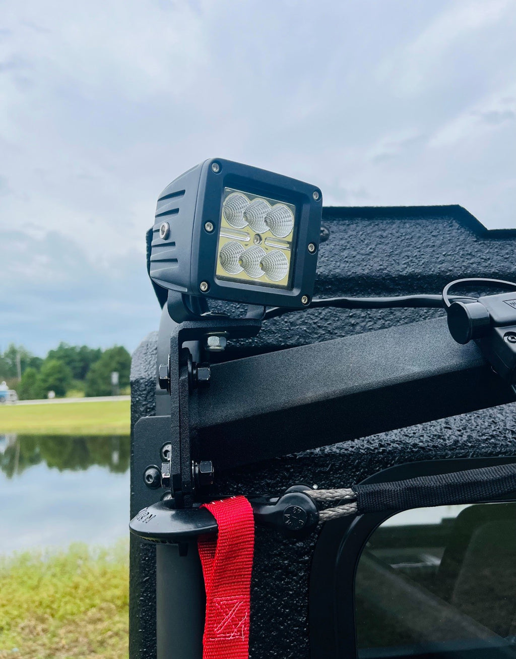 A Defender Game Loader is mounted near the roof and door of a rugged black vehicle, with a red fabric strap hanging below. In the background, grass, a pond, and a cloudy sky are visible.