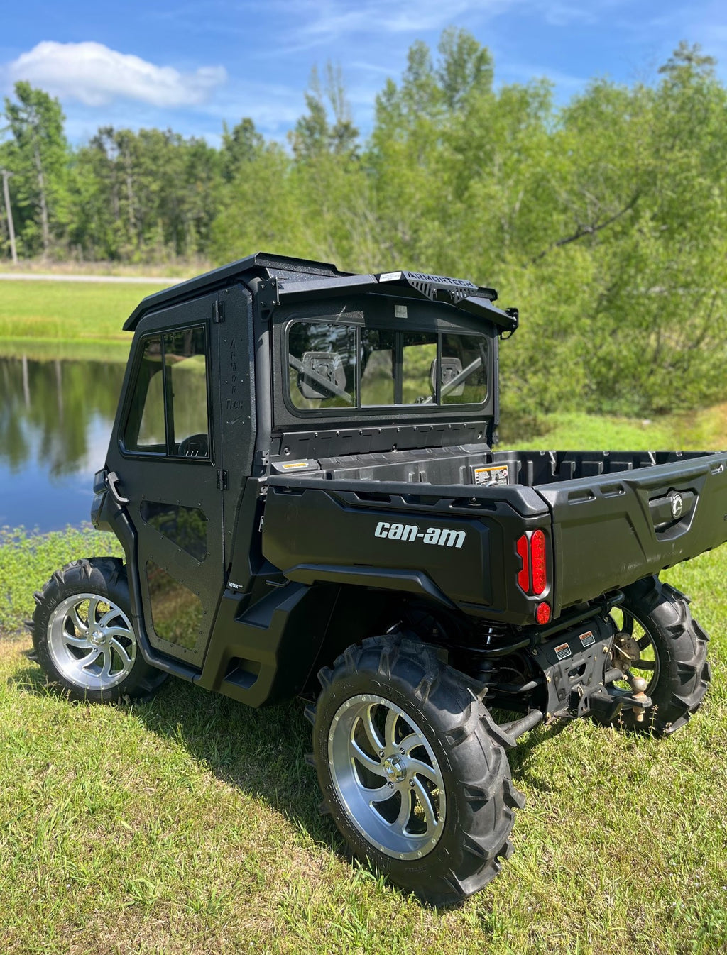 A black Can-Am Defender Game Loader side-by-side utility vehicle with large off-road tires, chrome rims, and a closed cab is parked on grass near a pond, with lush green trees and blue sky in the background.