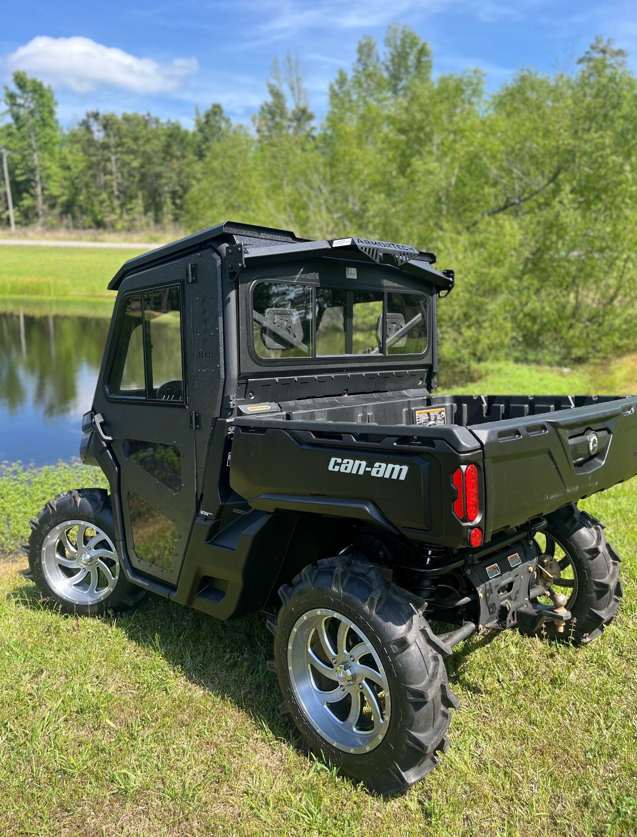 A black Can-Am Defender Game Loader side-by-side utility vehicle with large off-road tires, chrome rims, and a closed cab is parked on grass near a pond, with lush green trees and blue sky in the background.