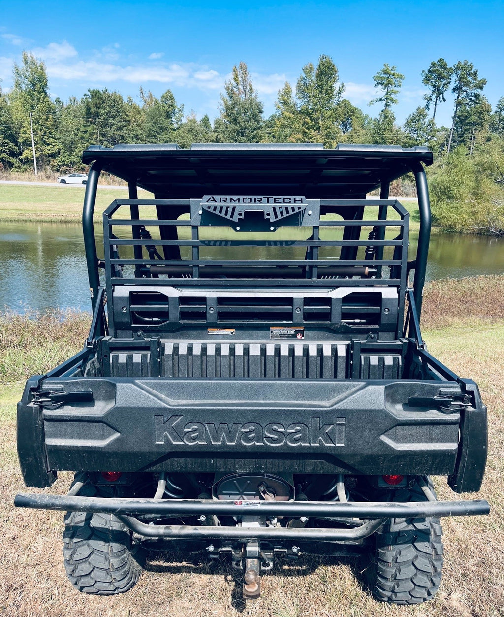 A rear view of a black Defender Game Loader utility vehicle parked on grass near a pond. It features a cargo bed, large off-road tires, rear metal rack, bumper, and branded logo. Trees and blue sky appear in the background.