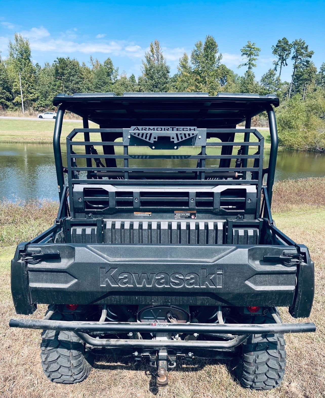 A rear view of a black Defender Game Loader utility vehicle parked on grass near a pond. It features a cargo bed, large off-road tires, rear metal rack, bumper, and branded logo. Trees and blue sky appear in the background.