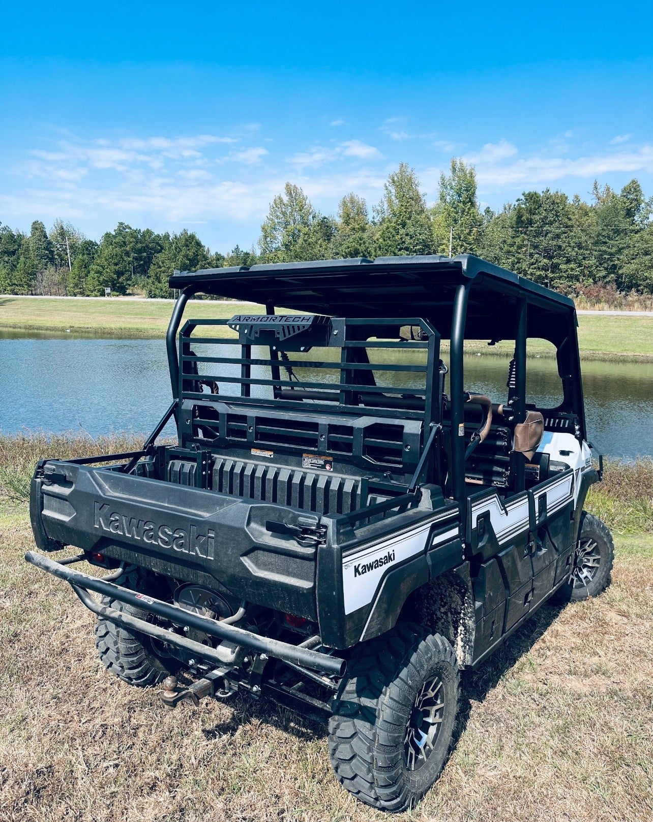 A Defender Game Loader utility vehicle in black is parked on dry grass near a small pond, facing away from the camera. Trees and shrubs border the far side of the pond beneath a bright blue sky with scattered clouds.