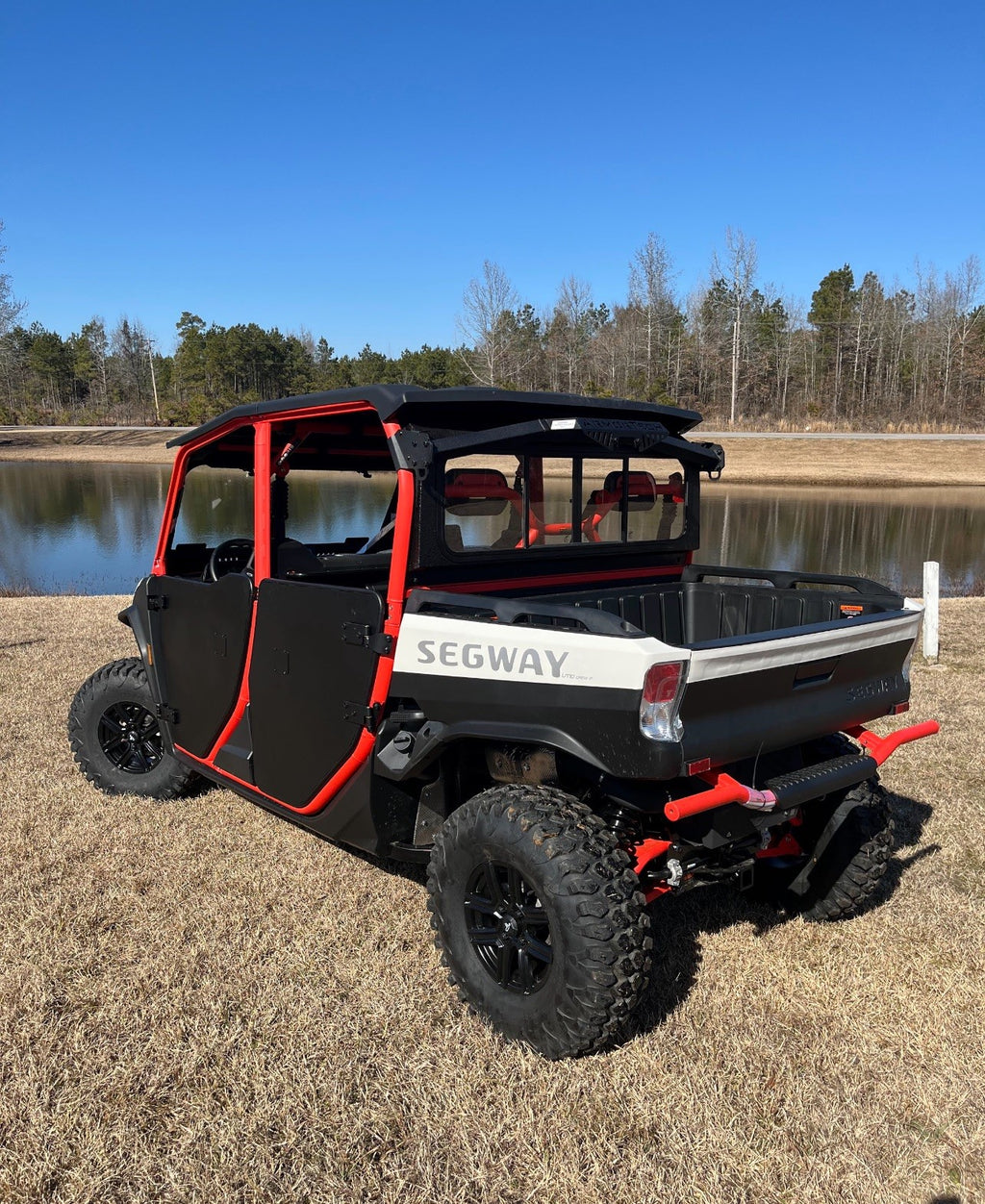 A black and red Defender Game Loader off-road utility vehicle with four doors and rugged tires is parked on dry grass near a calm pond, with leafless trees and a clear blue sky in the background.