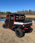 A black and red Defender Game Loader off-road utility vehicle with four doors and rugged tires is parked on dry grass near a calm pond, with leafless trees and a clear blue sky in the background.