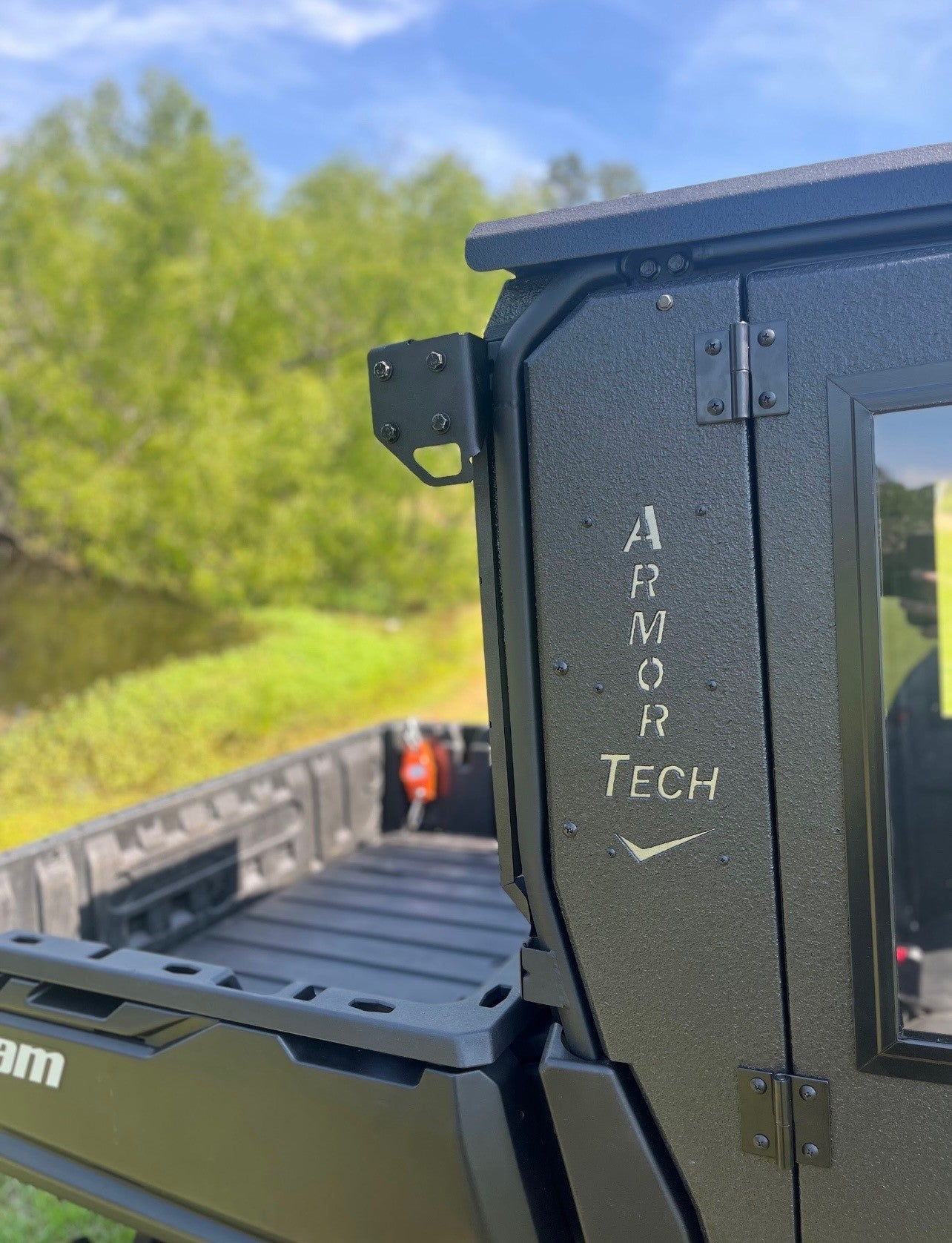 Close-up of a matte black Defender Game Loader with ARMOR TECH in white on the door. The truck bed and side mirror are visible, set outdoors with green trees and blue sky blurred in the background.