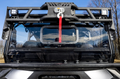 Close-up of a Polaris General Game Loader Rack mounted on a rugged utility vehicles front windshield, with a red-strap winch, black protective metal frame, and dual square LED lights. Leafless trees and blue sky with clouds in the background.