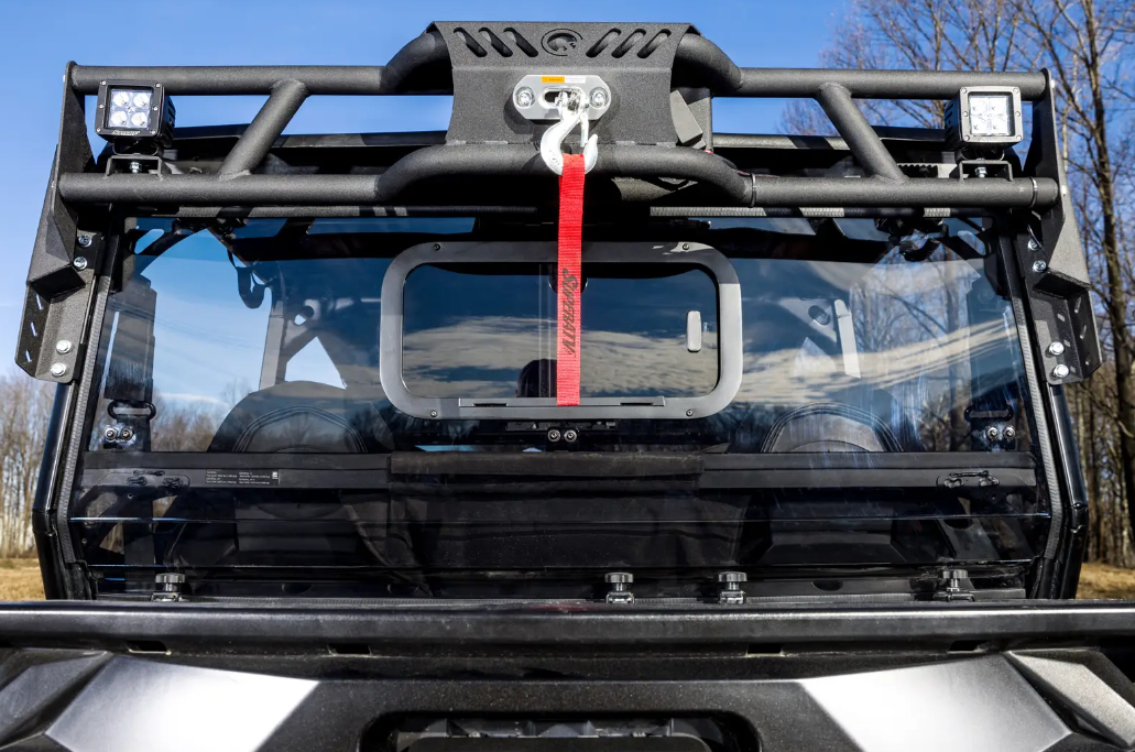 Close-up of a Polaris General Game Loader Rack mounted on a rugged utility vehicles front windshield, with a red-strap winch, black protective metal frame, and dual square LED lights. Leafless trees and blue sky with clouds in the background.