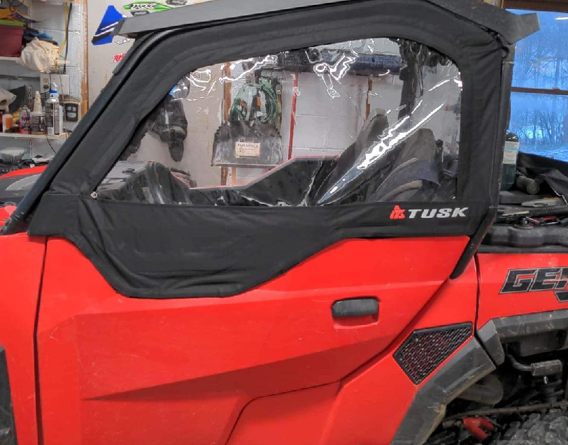 A red off-road vehicle with Polaris General Framed Upper Doors featuring clear windows is parked in a cluttered garage. Reflections, tools, shelves, and equipment are visible through the windows in the background.