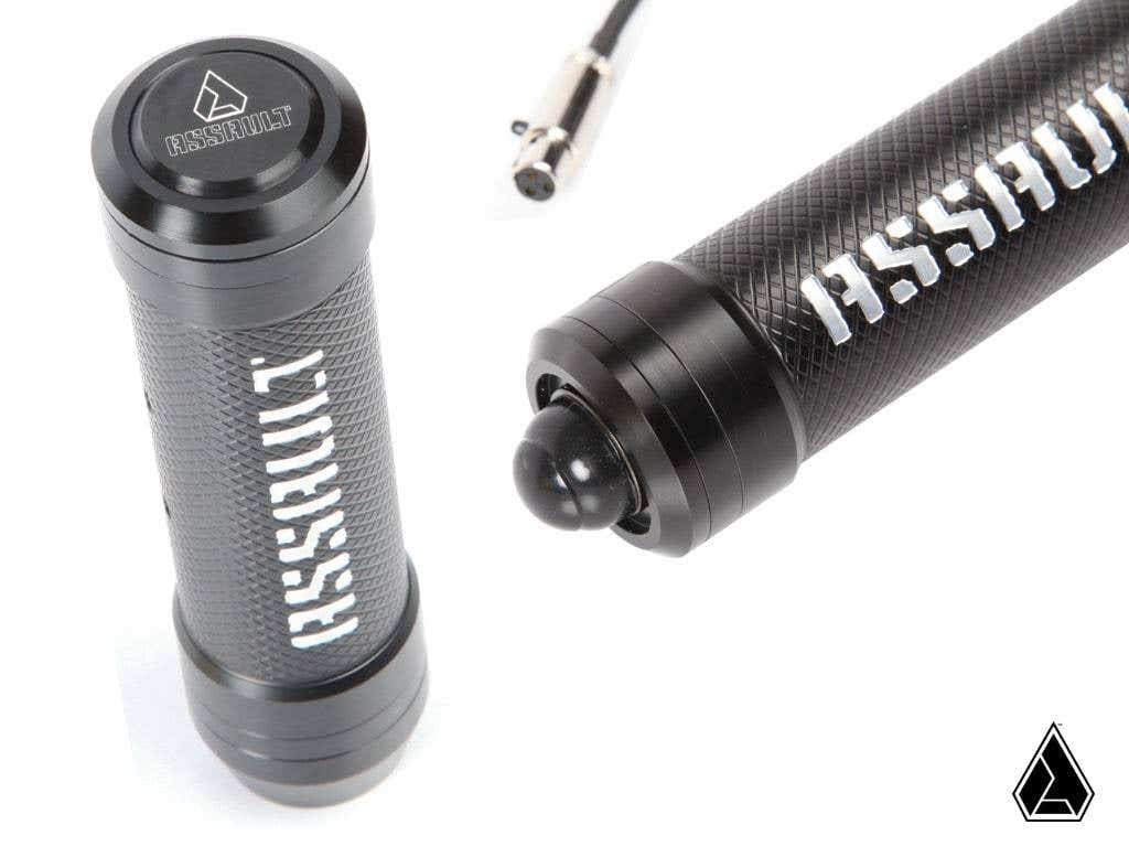 Close-up of two black Assault Industries M2HB Grab Handles for Can Am Maverick X3 with textured grips and bold white ASSAULT text. One shows a rounded button-like end, and a cable with connector is partially visible. Triangular logo at lower right.