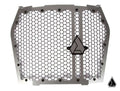 The Assault Industries Hellfire Grill for Kawasaki KRX 1000 is a silver metal grille with a hexagonal honeycomb pattern, thick frame, and triangular logos at the center and lower-right, designed for protection and ventilation.