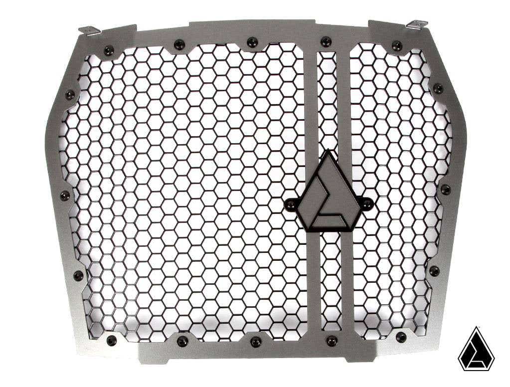 The Assault Industries Hellfire Grill for Kawasaki KRX 1000 is a silver metal grille with a hexagonal honeycomb pattern, thick frame, and triangular logos at the center and lower-right, designed for protection and ventilation.