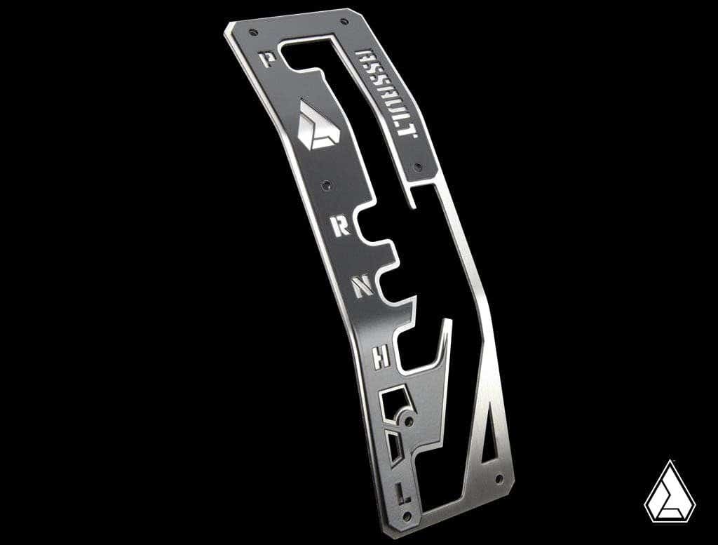 The Assault Industries L2R Shifter Gate Panel Kit for Can Am Maverick X3 features a metallic plate with cutouts for P, R, N, H, and L letters on a black background, a stylized logo, ASSAULT engraved at the top, and another small logo bottom right.