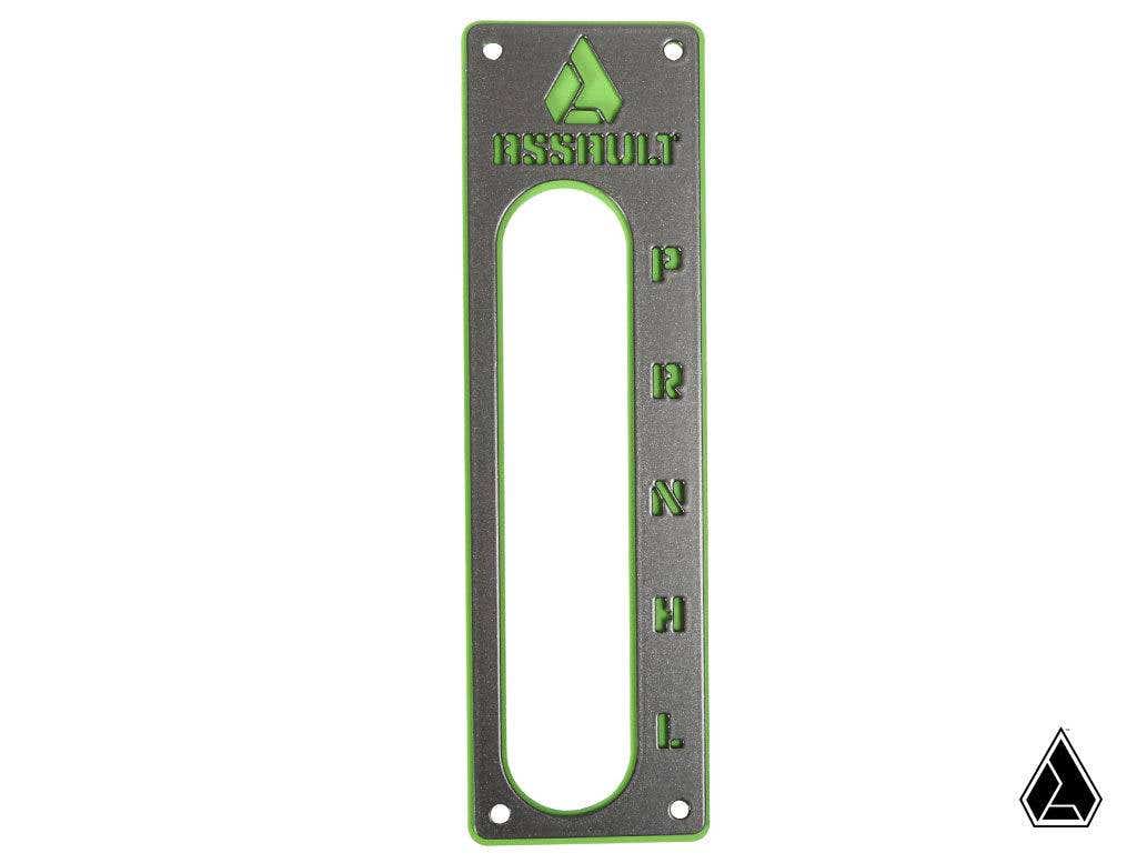 The Assault Industries Striker Shifter Gate Panel Kit for Wildcat XX/XTR1000 is a rectangular metallic gear shift plate with green accents, an elongated vertical cutout, P R N D L labels on the right, and Assault with a flame logo engraved at the top.