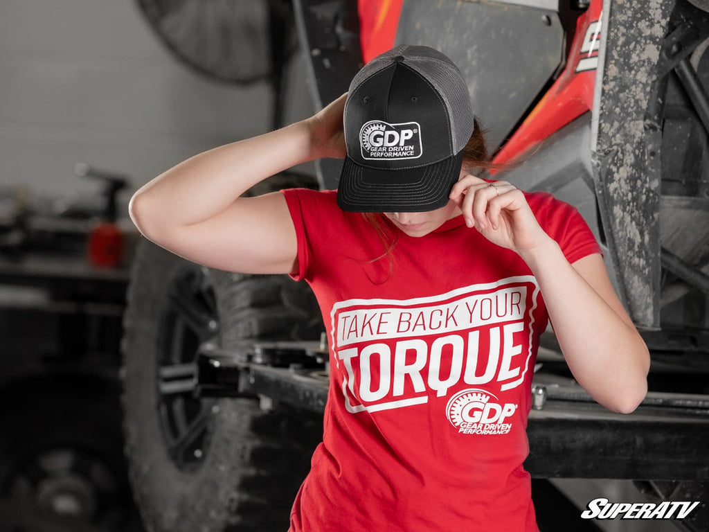 Wearing a red Take Back Your Torque t-shirt and the GDP Trucker Hat, a person stands before an off-road vehicle in a workshop, tilting the hat to partially hide their face. Vehicle parts and a blurred fan are visible in the background.