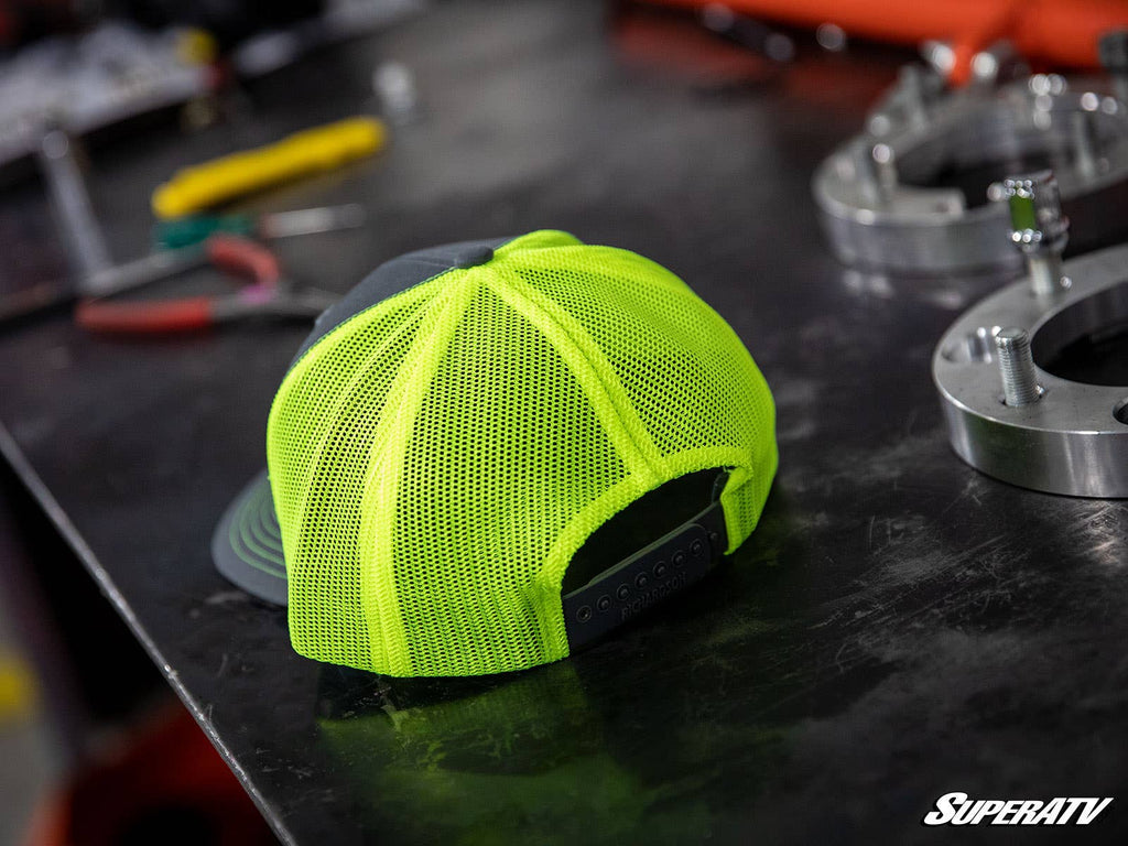 A green and gray SuperATV trucker hat sits on a metal workbench surrounded by tools and parts in a workshop. The SuperATV logo is visible in the bottom right corner.