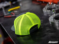 A green and gray SuperATV trucker hat sits on a metal workbench surrounded by tools and parts in a workshop. The SuperATV logo is visible in the bottom right corner.