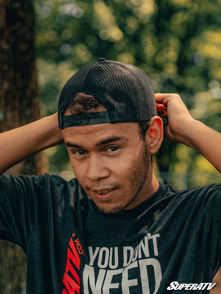 A young man with short hair and trimmed facial hair adjusts his black 15th Anniversary Trucker Hat. He wears a dark shirt reading YOU DON’T NEED in white text, with a sunlit forest behind him and the SuperATV logo in the lower right corner.