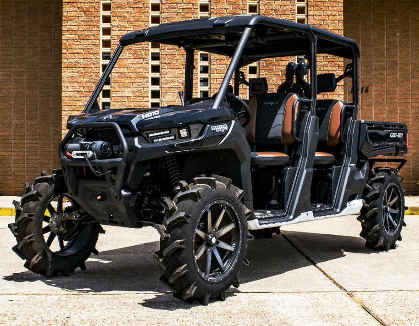 A rugged Can-Am Defender fitted with the S3 Power Sports 2016+ +2 Forward High Clearance A-Arm Kit, featuring large off-road tires, four brown seats, a roll cage, and cargo bed, parked on concrete by a tan brick wall.