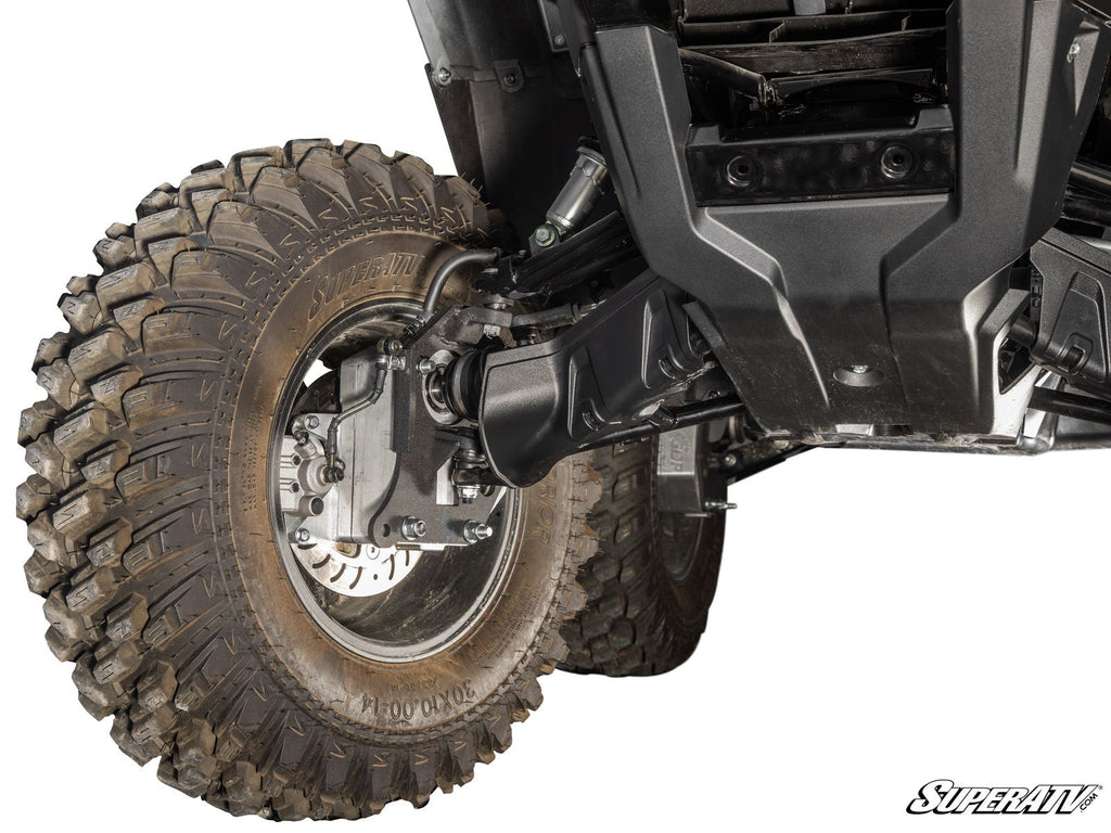Close-up of a Honda Rubicon 4 Portal Gear Lift installed, showing the front left wheel with a large, rugged tire, suspension parts, protective skid plate, and muddy tread. Detailed mechanical components and the SuperATV logo are visible in the corner.