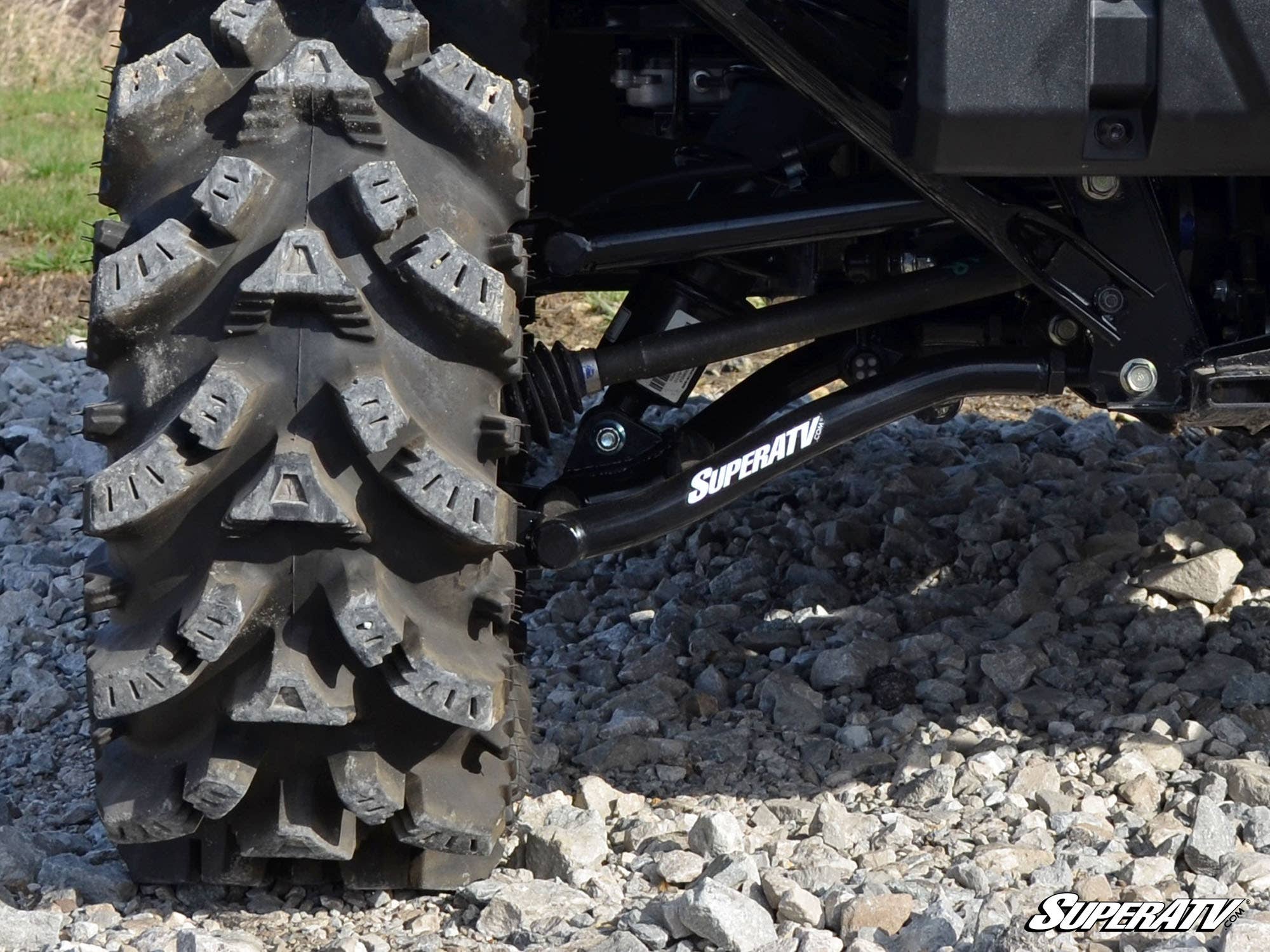 Close-up of a rugged off-road tire on rocky ground, with Honda Pioneer 1000 High Clearance 1.5 Offset Rear A-Arms featuring white SuperATV branding. Sunlight highlights the tire’s texture and rocks. SuperATV logo appears bottom right.